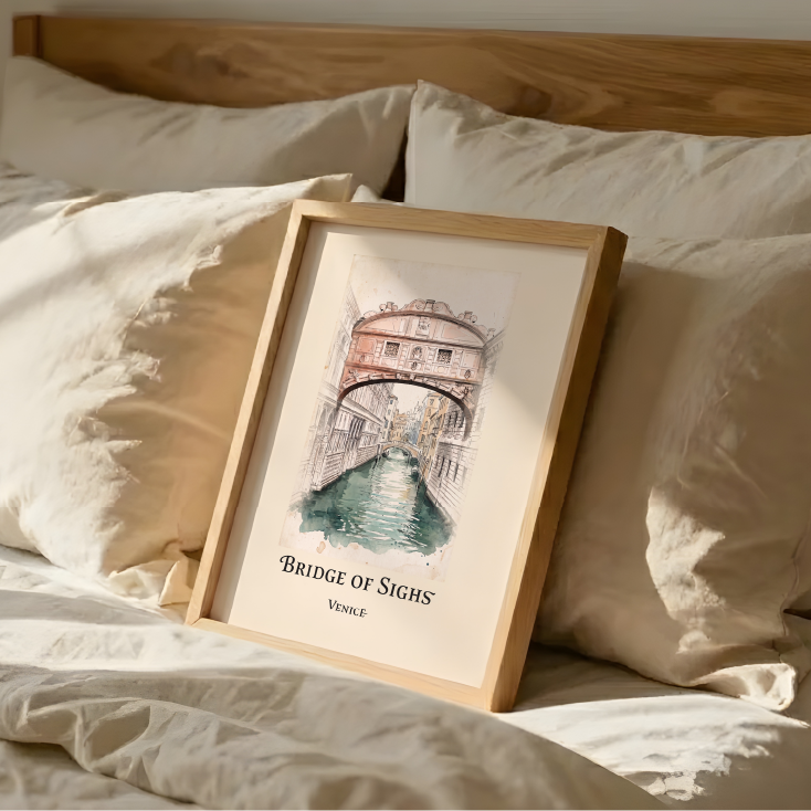 A framed art print featuring a watercolour illustration of the Bridge of Sighs in Venice, styled on a bed with beige bedding and pillows.