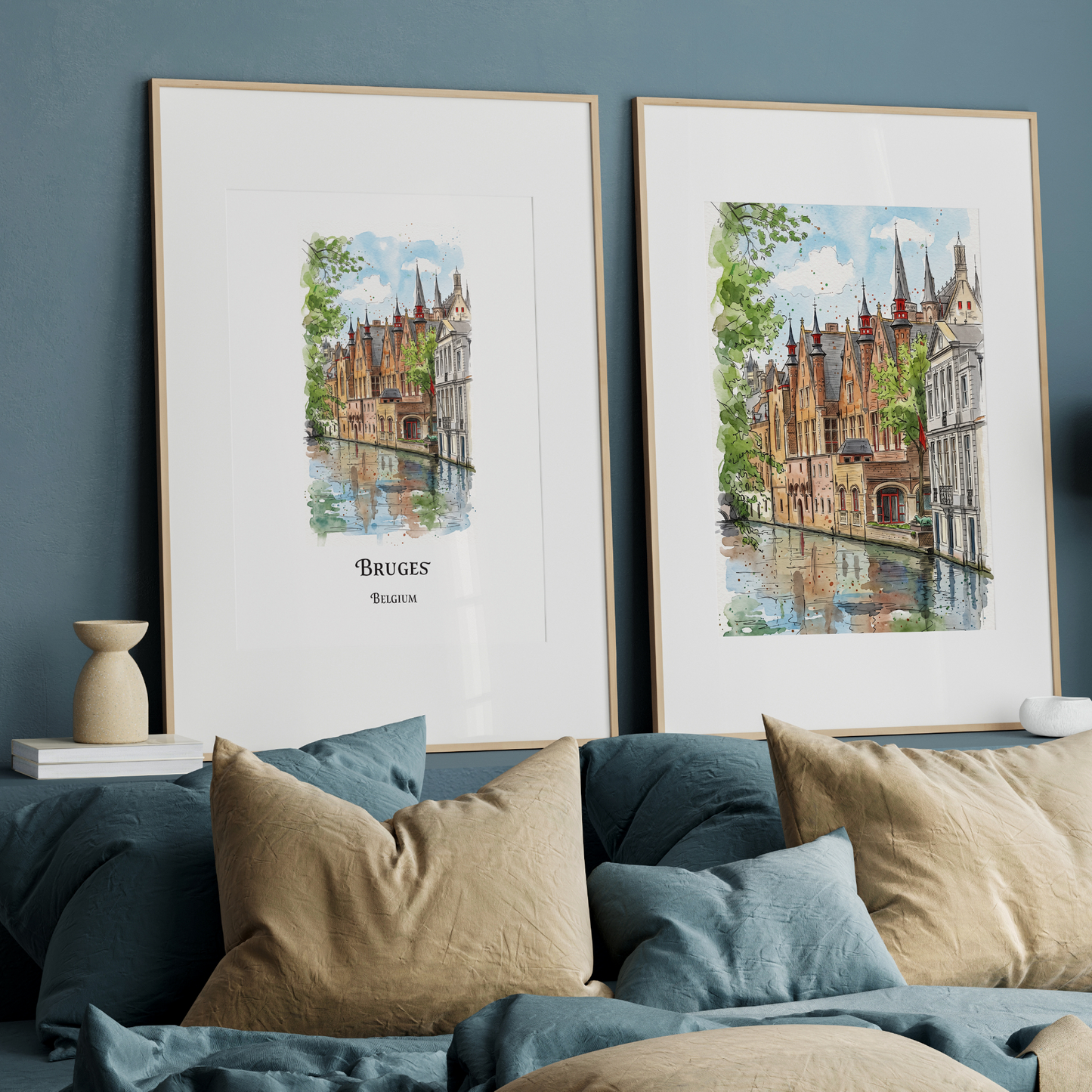 Two framed watercolour prints of Bruges, Belgium, displayed side-by-side on a dark blue wall above a bed with beige and blue pillows.