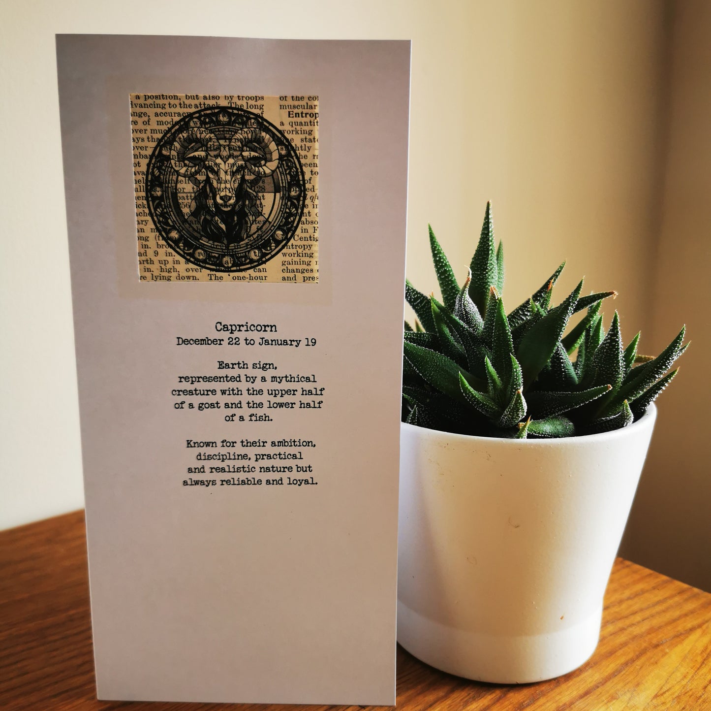 Card with Capricorn zodiac design and text next to a potted plant on a wooden surface.