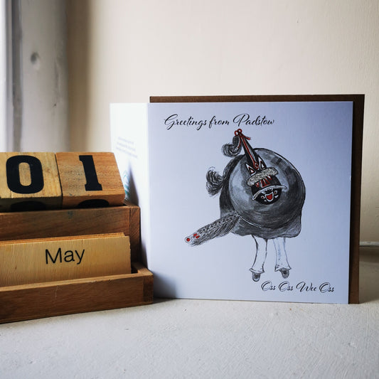 A greeting card with a watercolor illustration of a character named 'Red Obby Oss' accompanied by text 'Greetings from Padstow'. The card is placed next to a wooden block calendar showing '01 May'.