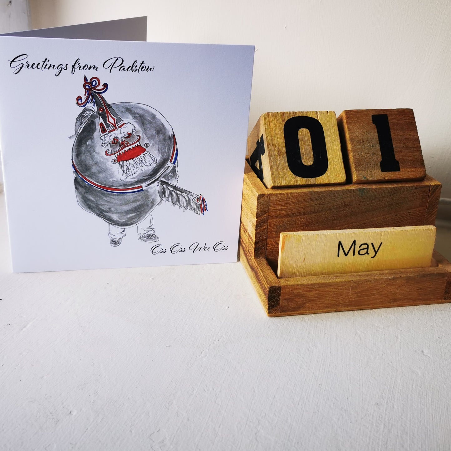 A greeting card featuring an illustration of a traditional Cornish 'Oss' character from Padstow, with a rustic brown envelope and a wooden block calendar showing '01' and the month 'May'.