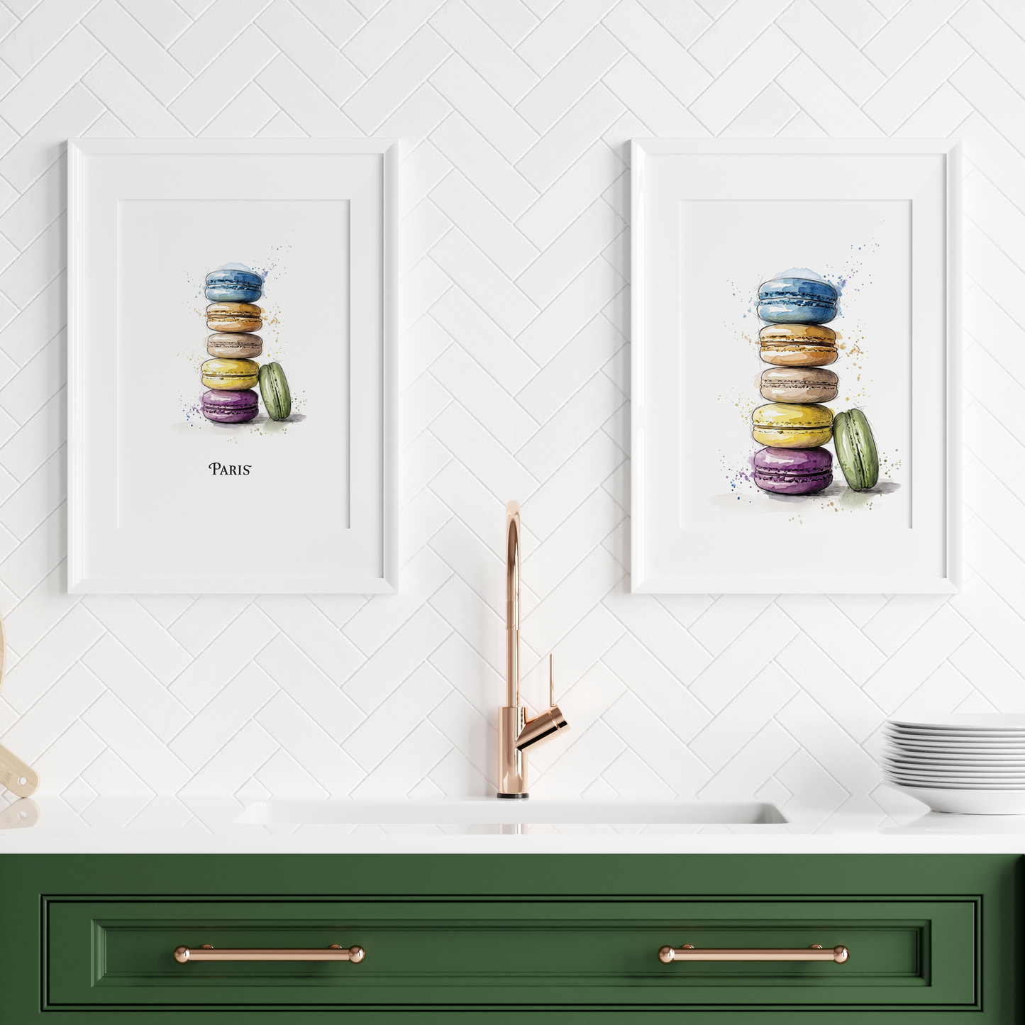 Two framed watercolour prints of stacked macarons hang on a white herringbone wall above a green kitchen counter with a faucet, plates, and a wooden board.