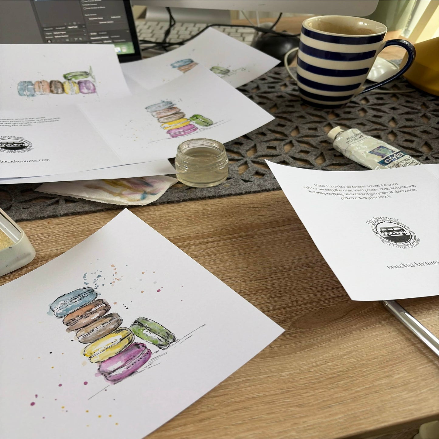 A wooden desk with hand painted artwork greeting cards showing colourful macaron illustrations, a blue striped ceramic mug, a small jar, and other desk items in a real indoor setting.