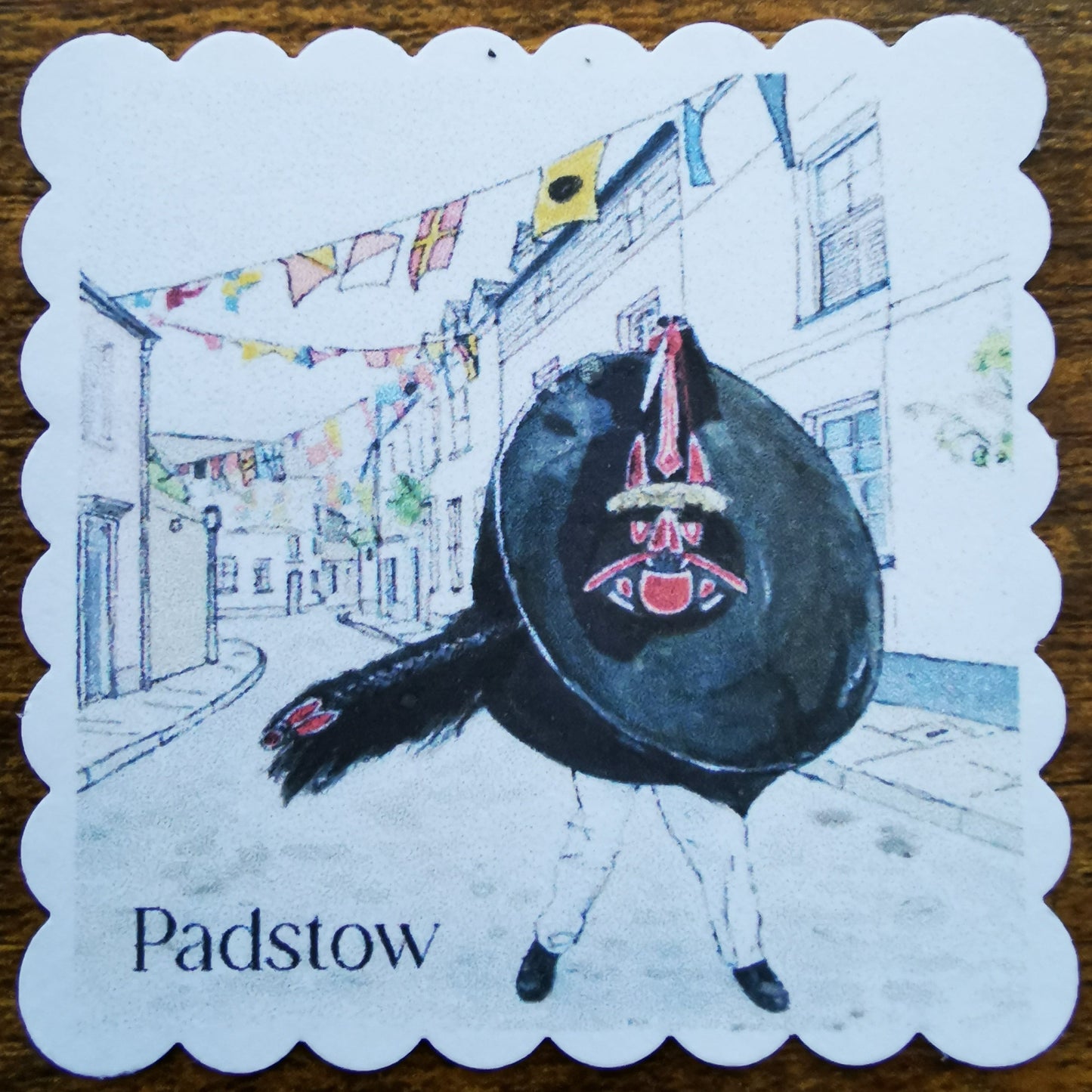Square faux postage stamp with a design of a Red Obby Oss in a street scene and 'Padstow' text on a wooden surface