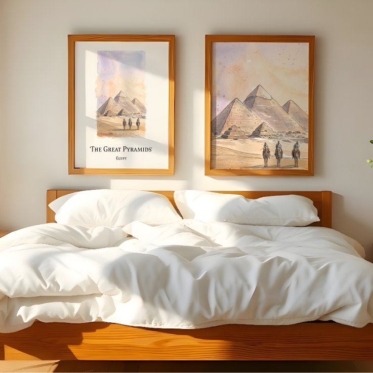 A styled bedroom scene with a wooden bed dressed in white bedding, flanked by matching nightstands and a lamp, with two framed wall art prints above the headboard showing desert pyramids.