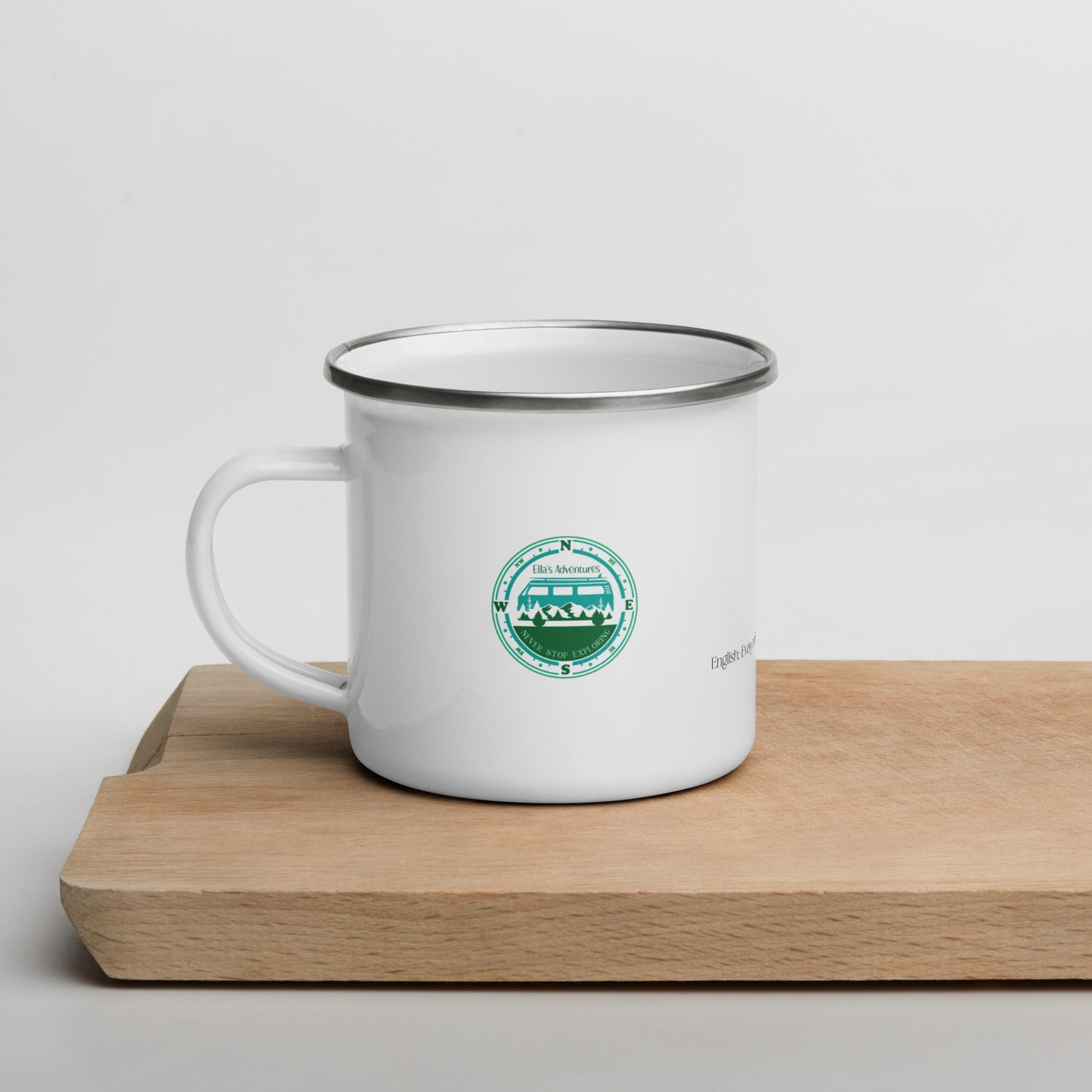 white enamel mug with Ellas Adventure logo in blue an dgreen on a wooden chopping board