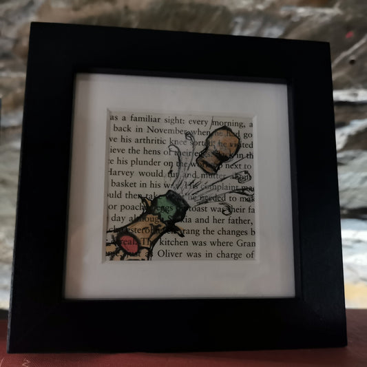 A freestanding black 12.5 cm box frame on a bookshelf against a back lit slate wall. An internal 6cm frame has a book page with a hand-drawn watercolour illustration of a champagne bottle pointing up to the right with the cork popping.