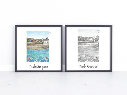 Two framed art prints of Bude Seapool, one in a watercolour style with colourful beach huts and blue water, and one in a black and white sketch style, displayed side-by-side on a white marble surface against a white wall.