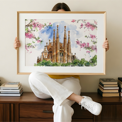 A person sitting on a wooden cabinet holds up a large framed watercolour painting of Sagrada Familia in Barcelona, with pink blossoms framing the scene. The room has a beige wall and stacks of books on either side.