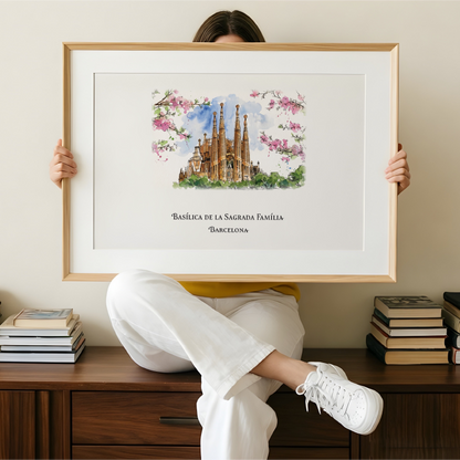 A person sitting on a wooden cabinet holds up a large framed art print featuring a watercolour illustration of the Sagrada Familia basilica in Barcelona, with pink flowers and the text 'Basílica de la Sagrada Familia Barcelona' below. The room has a beige wall, and stacks of books are placed on either side of the cabinet.