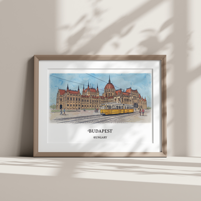 A framed watercolor art print of Budapest, Hungary, featuring the Hungarian Parliament Building and a yellow tram, displayed on a shelf against a wall with soft leaf shadows.