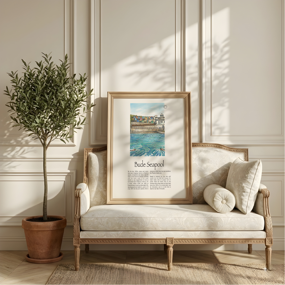 A framed art print featuring a colourful illustration of Bude seapool with the text 'Bude Seapool' and descriptive text below, styled on a cream-coloured wooden chaise lounge in a bright, elegant room next to a potted olive tree.