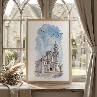 A framed watercolor art print of Caracas cathedral, Venezuela under a blue sky, leaning on a windowsill in a softly lit room with arched windows, beige curtains, and dried grasses in the foreground
