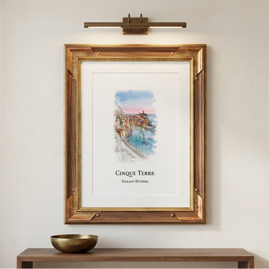 A framed art print featuring a watercolour painting of Cinque Terre, Italian Riviera, displayed on a light beige wall above a wooden console table with a brass bowl. A modern wall-mounted brass light fixture is positioned above the frame.