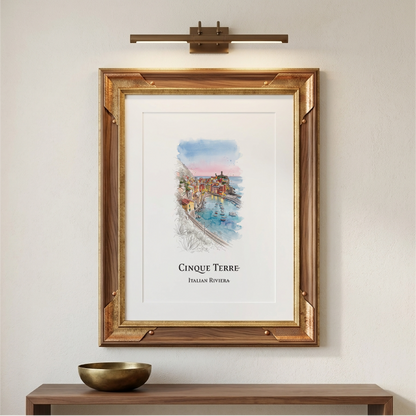 A framed art print featuring a watercolour painting of Cinque Terre, Italian Riviera, displayed on a light beige wall above a wooden console table with a brass bowl. A modern wall-mounted brass light fixture is positioned above the frame.