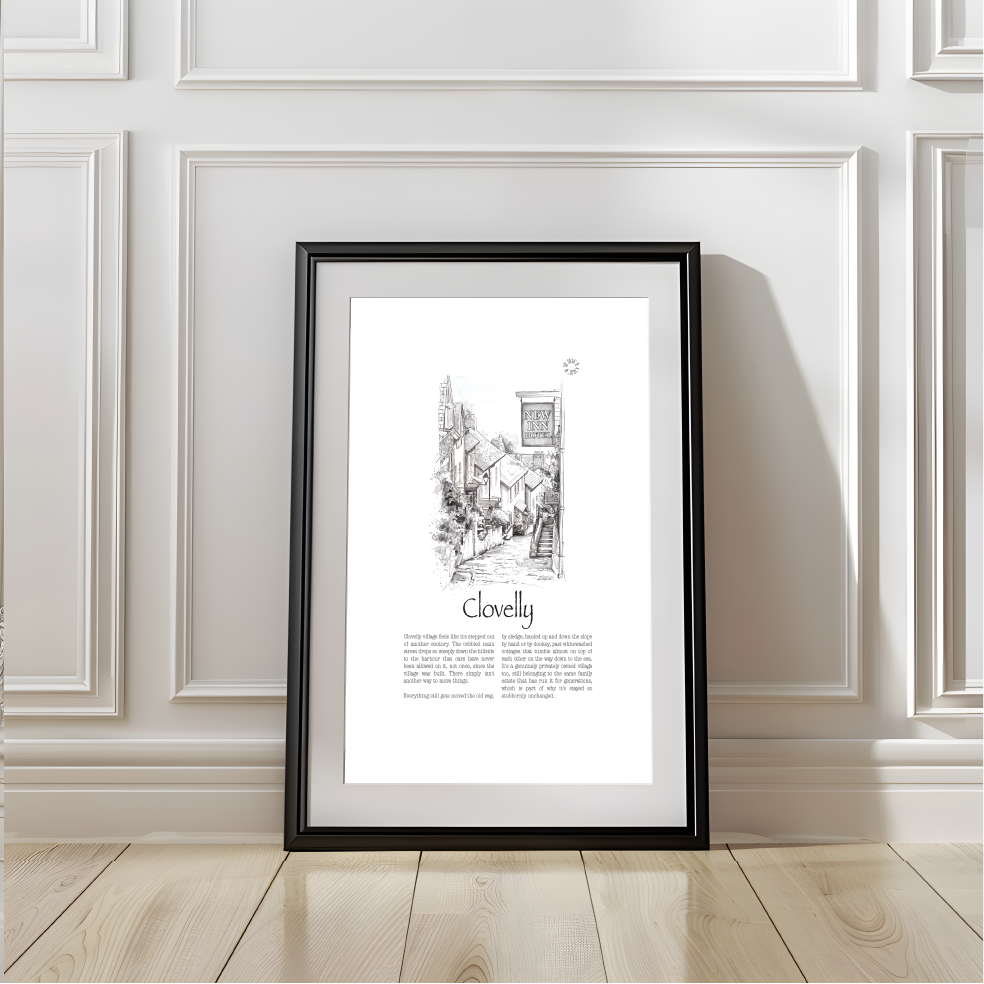 A framed art print leaning against a white wood pannelled wall in a sunlit living room. The print features a black and white illustration of a quaint village street with the text 'New Inn' and 'Clovelly', surrounded by a black wood frame.