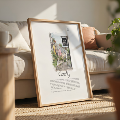 A framed art print leaning against a beige sofa in a sunlit living room. The print features a watercolour illustration of a quaint village street with the text 'New Inn' and 'Clovellly', surrounded by a light wood frame.