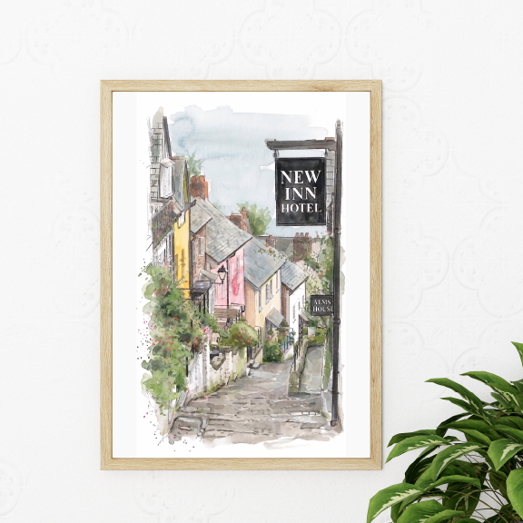 A framed watercolour art print depicting a quaint street scene in Clovelly, Devon, with the 'New Inn Hotel' sign, styled on a white wall next to a green houseplant.