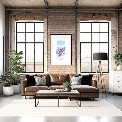 A styled living room interior with a brown leather sofa, black accent pillows, a black coffee table, brick walls, large industrial windows, potted plants, and a framed print of Colca Canyons hanging above the sofa.