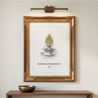 A framed art print featuring a gold-coloured chocolate museum chocolate fountain on a white background, styled on a beige wall above a wooden console table with a gold bowl. A brass wall sconce is mounted above the frame.