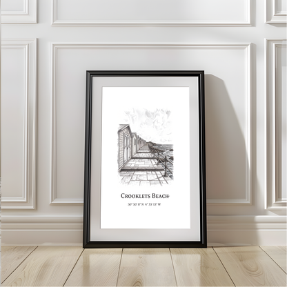 A framed black and white sketch poster of Crooklets Beach, displayed leaning against a white paneled wall on a light wood floor.