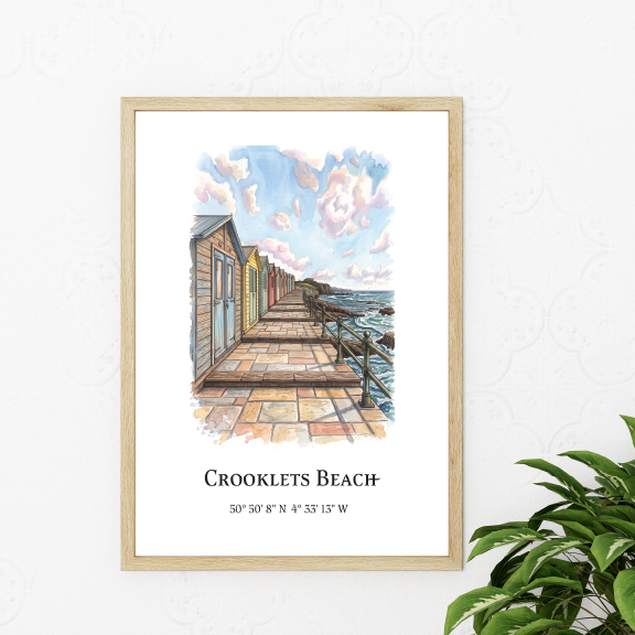 A framed art print depicting a watercolour illustration of Crooklets Beach with a boardwalk, beach huts, and ocean view, styled on a white wall next to a green plant.