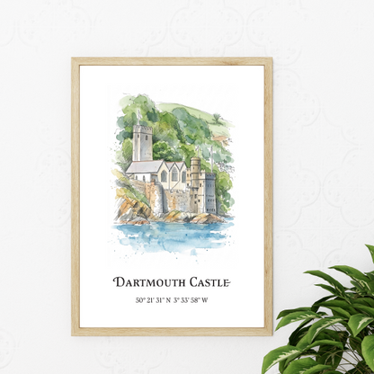 A framed watercolour art print of Dartmouth Castle, featuring a light wood frame and a white background with the title and coordinates printed below. The print is styled on a white wall next to a green houseplant.