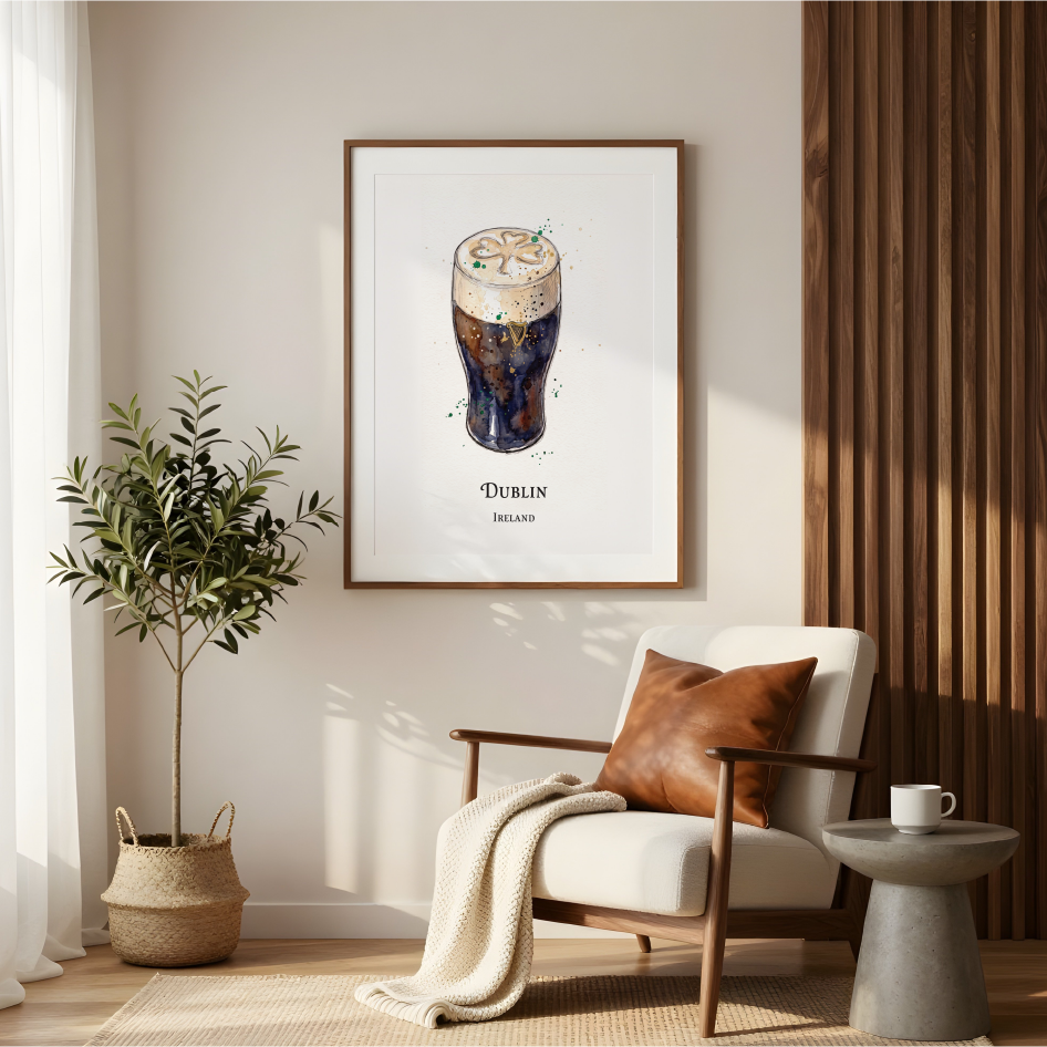 A framed art print featuring a vintage watercolour illustration of a foamy dark guiness in a pint beer glass with the text 'DUBLIN' and 'Ireland' below, displayed on a beige wall in a modern living room setting with a chair, plant, and side table.