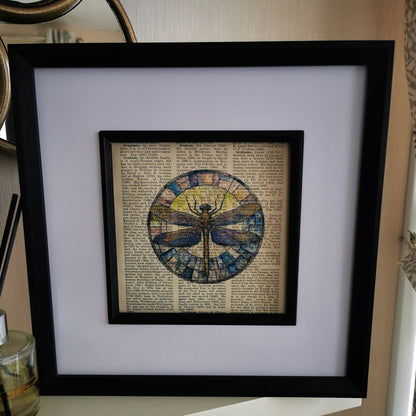A watercolour and ink illustration of a dragonfly, crafted on a century-old encyclopaedia page, with visible text and aged patina photograped on a wooden shelf