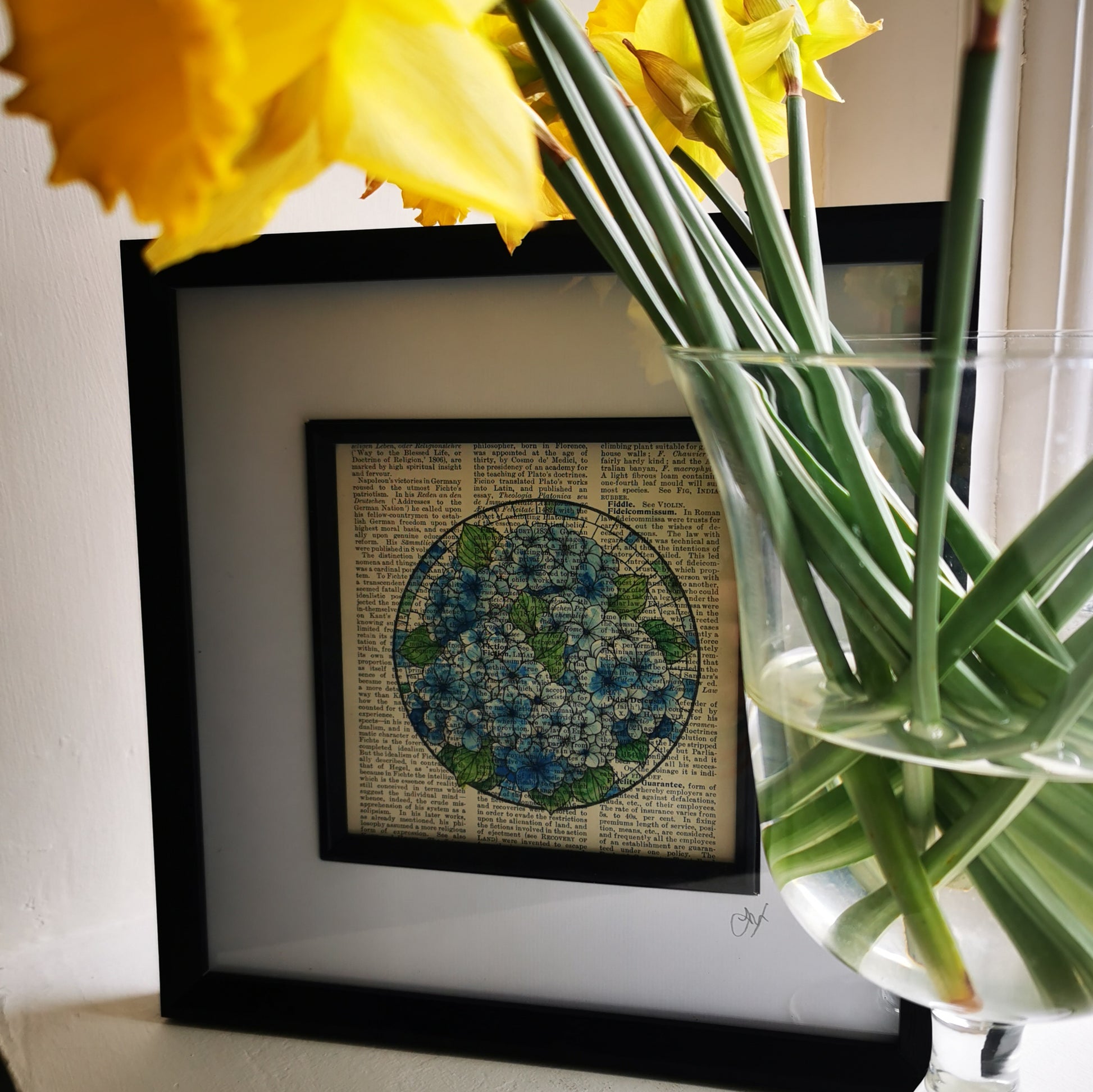 A framed art piece featuring a watercolour and ink illustration of blue hydrangea flowers on a vintage book page, displayed on a windowsill with daffodils in the foreground.