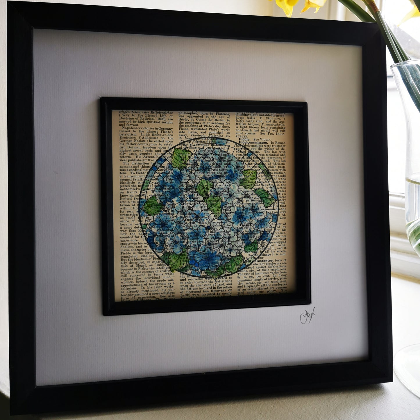 A framed art piece featuring a watercolour and ink illustration of blue hydrangea flowers on a vintage book page, displayed on a windowsill with daffodils in the background.
