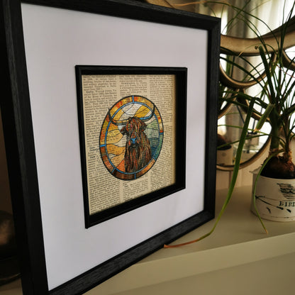 Framed artwork featuring a watercolour and ink illustration of a Highland Cow on a vintage encyclopaedia page, displayed on a shelf with a plant in the background.