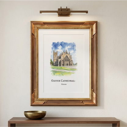 A framed art print of Exeter Cathedral, featuring a watercolour painting of the cathedral under a blue sky, displayed on a beige wall above a wooden console table with a gold bowl. A brass wall sconce is mounted above the frame.