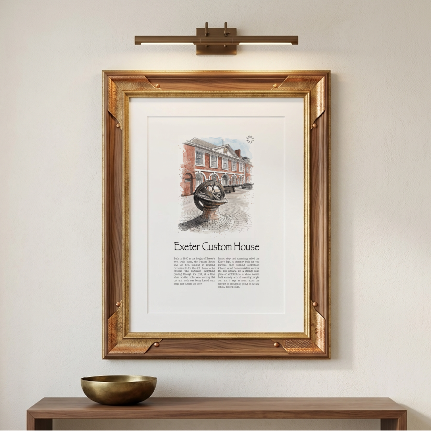 A framed art print featuring a vintage watercolour illustration of Exeter Custom House with text below, displayed on a beige wall above a wooden console table with a brass bowl. A modern brass wall sconce is mounted above the frame.