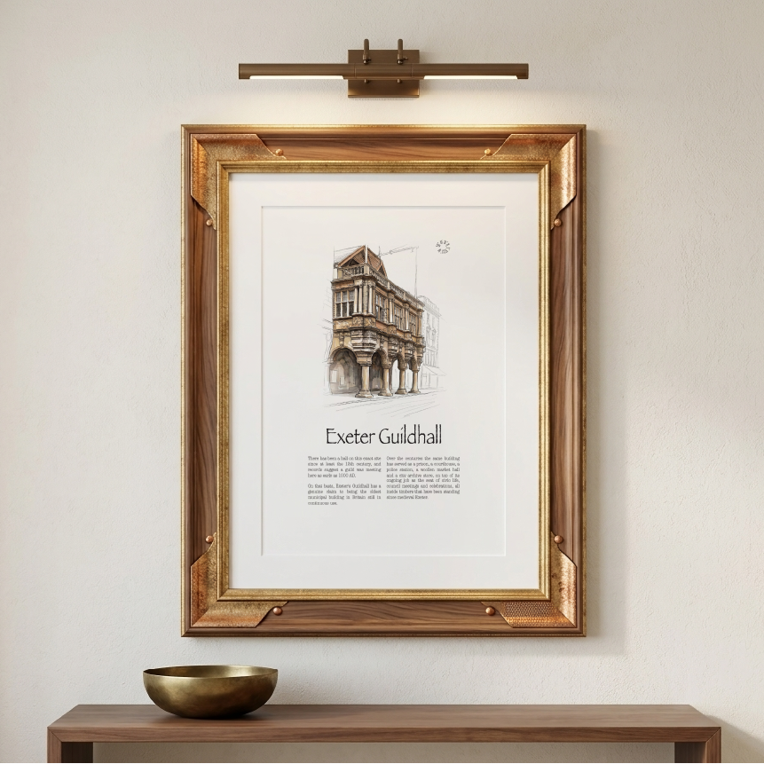 A framed art print featuring a watercolour sketch of Exeter Guildhall, displayed on a beige wall above a wooden console table with a brass bowl. A modern brass wall sconce is mounted above the frame.