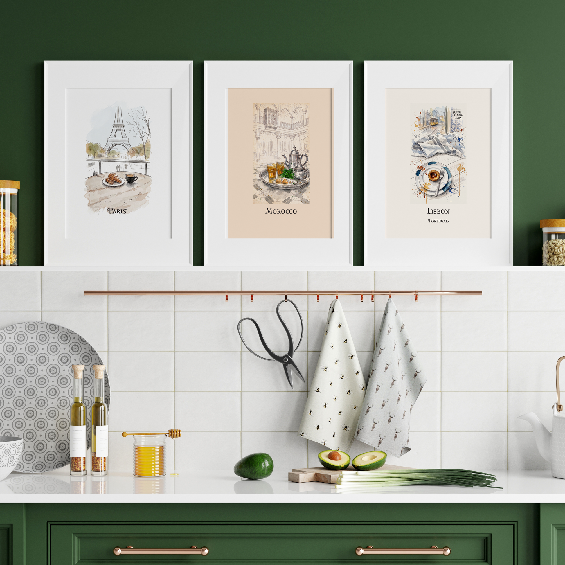 Three framed art prints featuring Paris, Morocco, and Lisbon illustrations, displayed on a dark green wall above a white tiled kitchen counter with green cabinets and copper-toned handles.