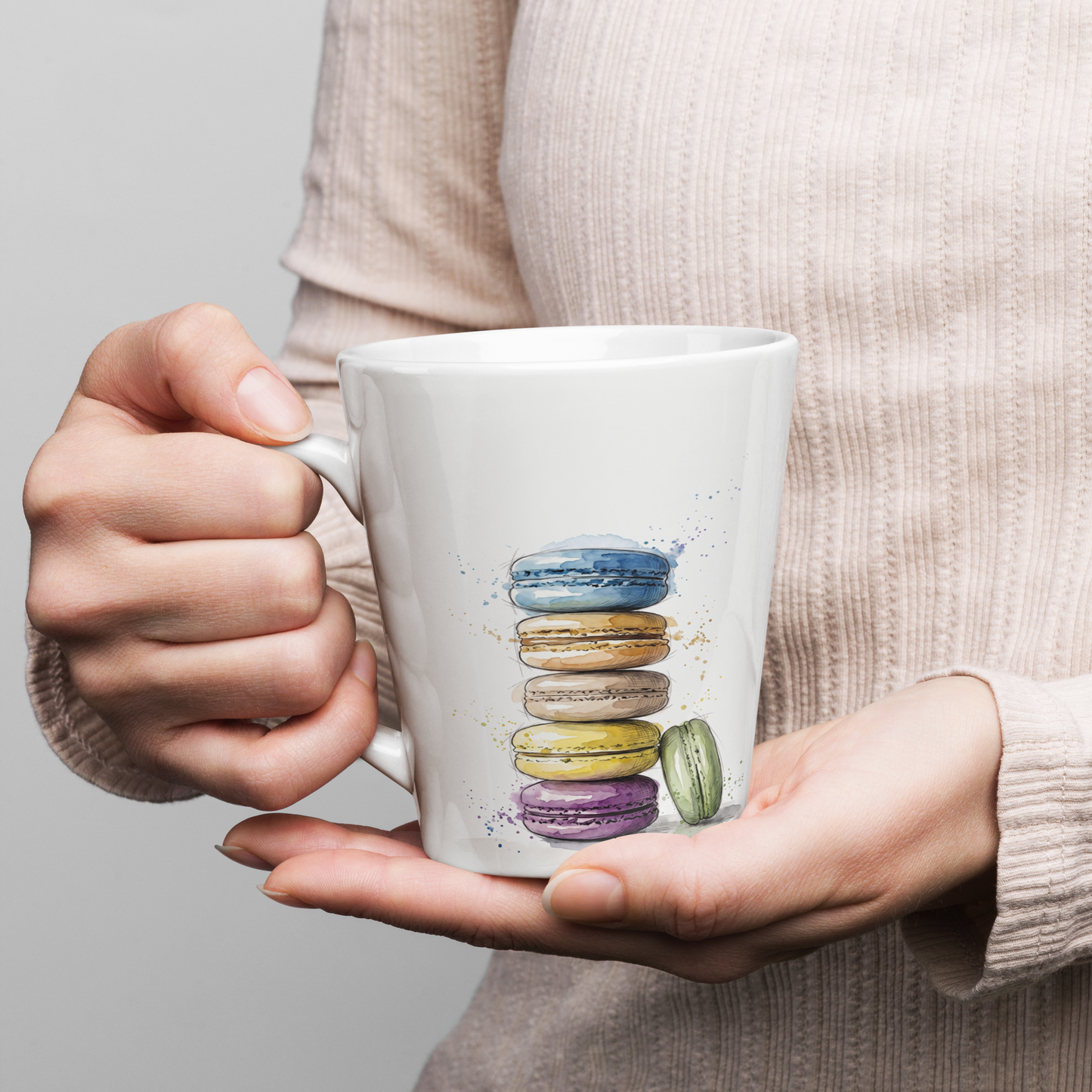 A person in a beige knit sweater holds a white ceramic mug featuring a colourful watercolour illustration of stacked macarons.