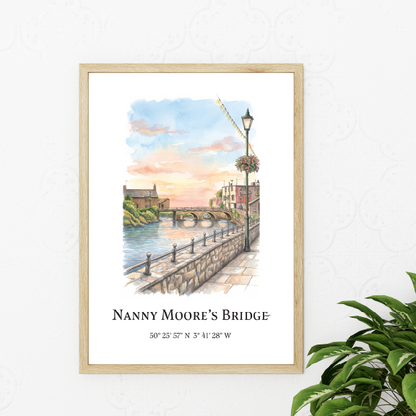A framed watercolour art print of Nanny Moore's Bridge in Bude, Cornwall, displayed on a white wall next to a green potted plant. The print features a light wood frame and includes the title and coordinates at the bottom.