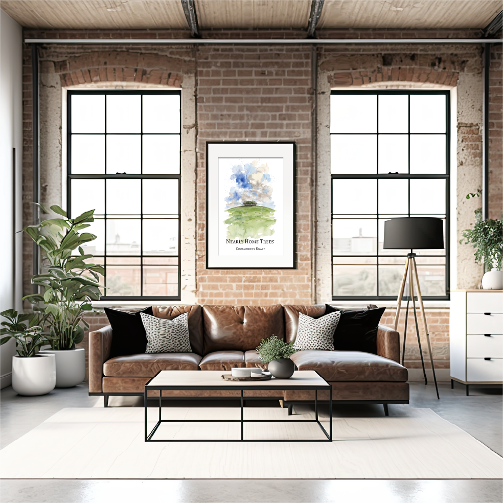 A styled living room featuring a brown leather sofa with black and white patterned pillows, a black metal coffee table, a white rug, brick walls, and two large industrial windows. A framed art watercolour print  of The Nearly Home Trees on the Devon/Cornwall border hangs on the wall above the sofa.