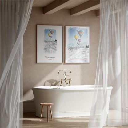 A styled bathroom scene with two framed art prints featuring Pamukkale Travel Posters with hot air balloons over snowy landscapes, hung above a white freestanding bathtub with a brass faucet. White sheer curtains frame the scene, and a small wooden stool sits beside the tub.