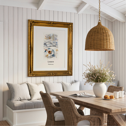 A framed art print featuring a watercolour illustration of Pastel de Nata tarts with the text 'LISBON PORTUGAL' hangs on a white shiplap wall above a dining nook with striped cushions. The print is housed in an ornate gold frame and is styled in a bright, modern dining room with a wooden table, wicker chairs, and a woven pendant light.