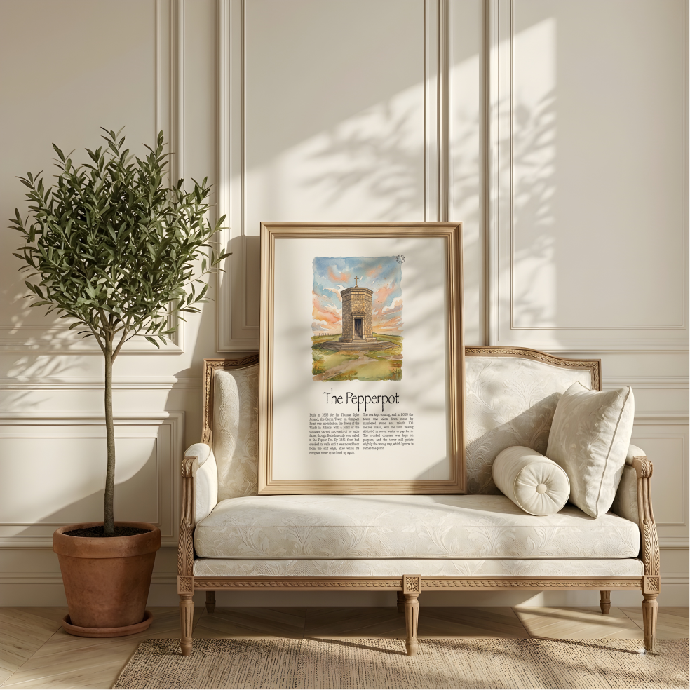 A framed art print titled 'The Pepperpot' leaning against a cream-coloured upholstered wooden chaise lounge in a styled room with a potted olive tree and warm sunlight.