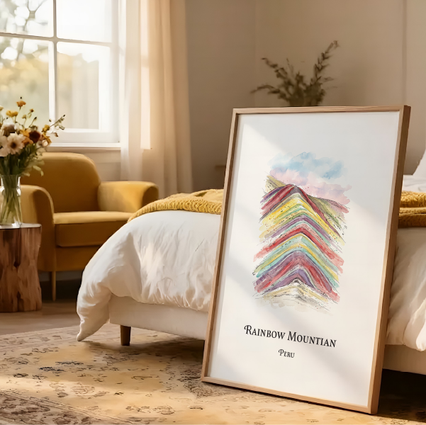 A framed art print leaning against a bed in a warm, modern bedroom. The print features a colourful, rainbow-striped mountain design with the text 'RAINBOW MOUNTAIN' and 'Peru' below it.