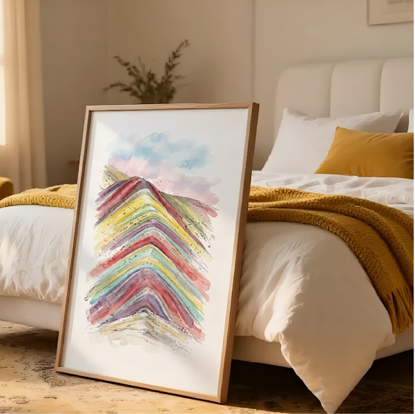 A framed abstract watercolour art print leaning against a white bed in a warm, modern bedroom setting. The print features layered, colorful chevron-like mountain in Peru, in shades of red, yellow, green, blue, and purple on a white background, enclosed in a light wood frame.