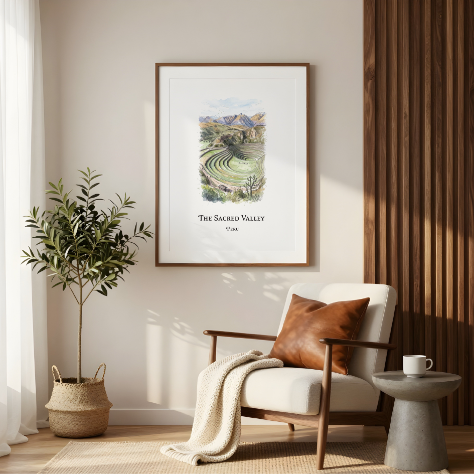 A framed art print titled 'The Sacred Valley' hanging on a beige wall in a modern living room setting with a white armchair, a potted plant, and a side table.