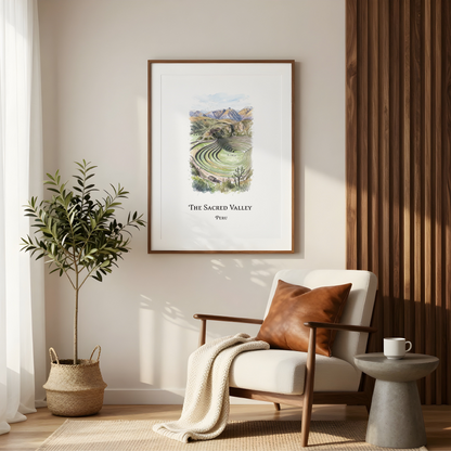 A framed art print titled 'The Sacred Valley' hanging on a beige wall in a modern living room setting with a white armchair, a potted plant, and a side table.