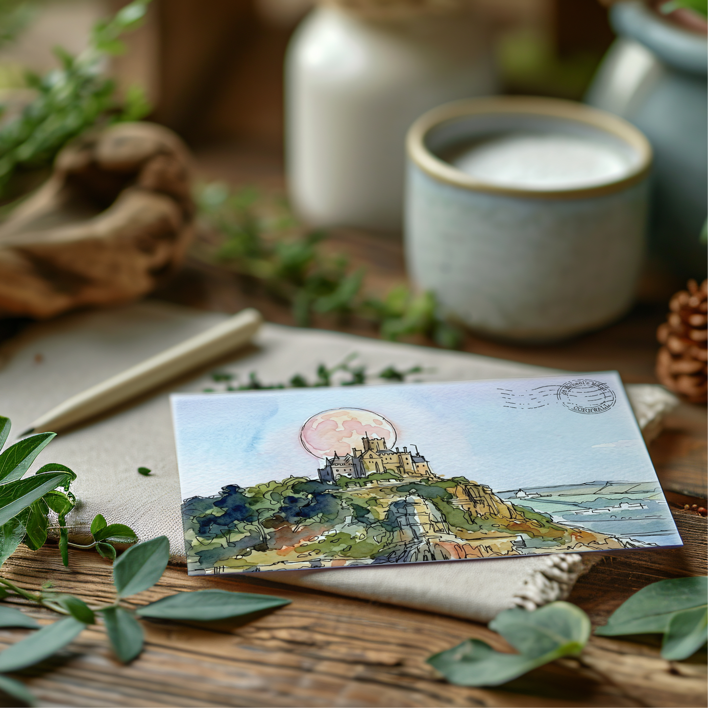 A rectangular art print or postcard featuring a watercolour landscape of St Michaels Mount with a large pink moon, styled on a wooden surface with leaves, a candle, and ceramic vessels in the background.