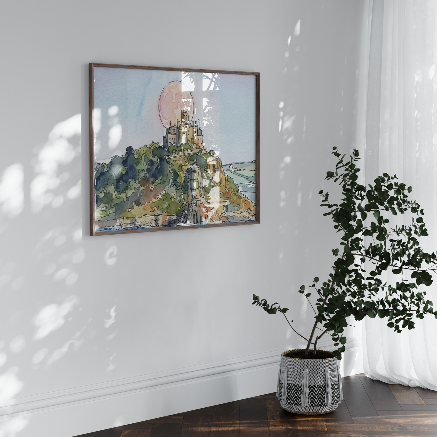 A framed watercolor painting of a St Michaels Mount castle displayed on a white wall in a styled room setting with a potted plant on a wooden floor and soft window light casting shadows.