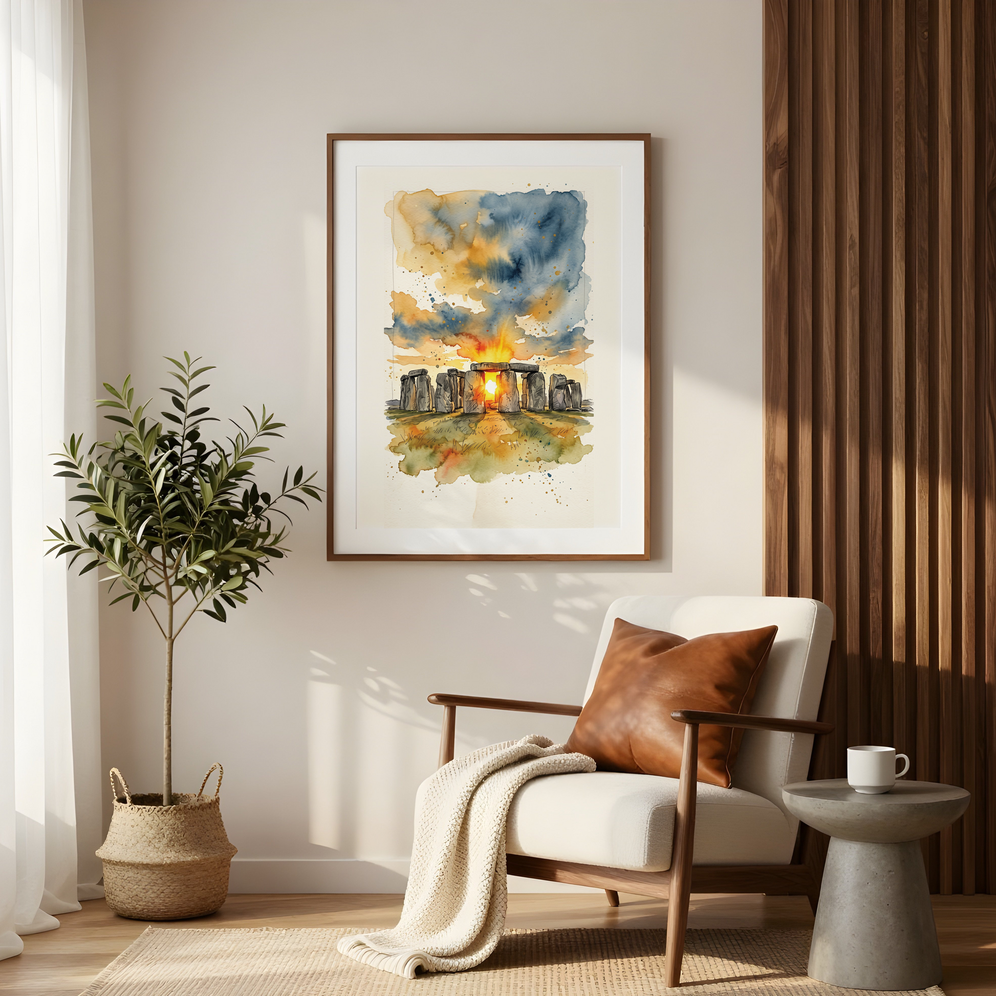 A framed watercolour art print of Stonehenge at sunset hangs on a beige wall in a modern, sunlit living room with a white armchair, a potted plant, and a small side table.