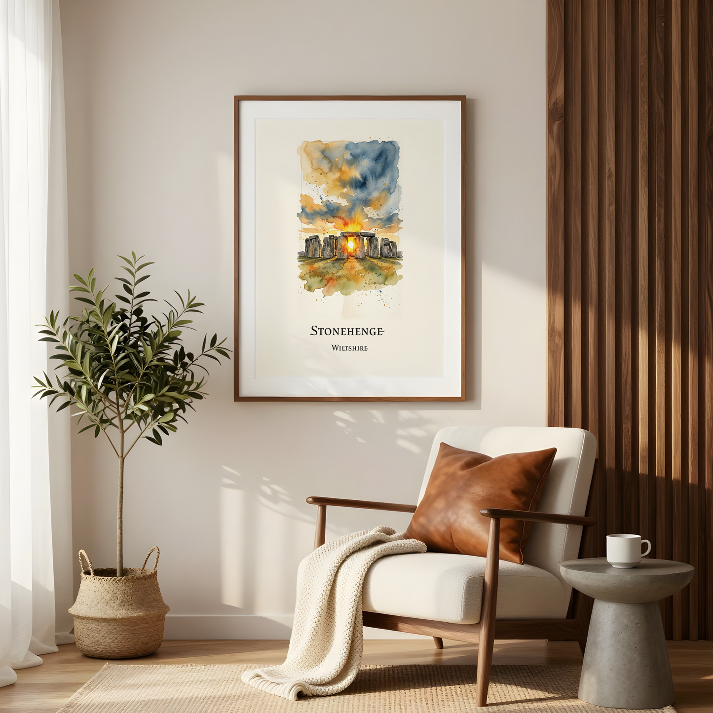 A framed watercolour art print of Stonehenge, Wiltshire hanging on a beige wall in a modern, sunlit living room with a white armchair, a potted plant, and a side table.